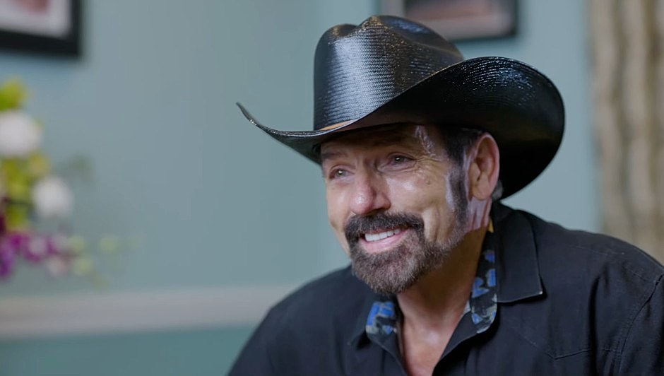 Man in a cowboy hat smiling warmly indoors.